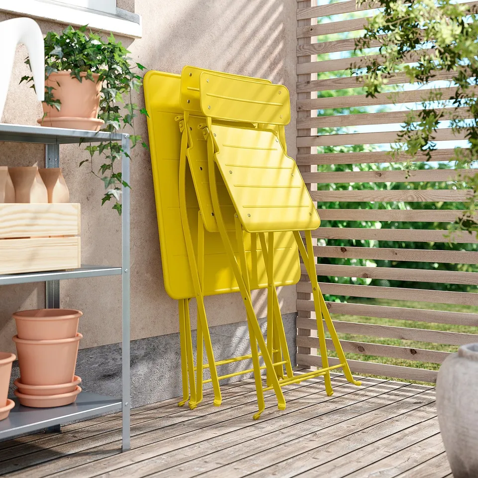 Three yellow metal chairs stacked against a wooden fence on a balcony with potted plants nearby.