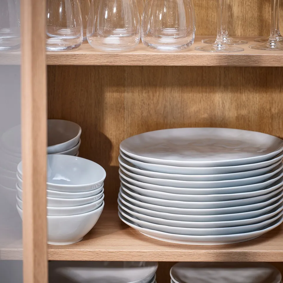 Shelves filled with stacks of white porcelain plates, bowls, and clear glassware, neatly organised.