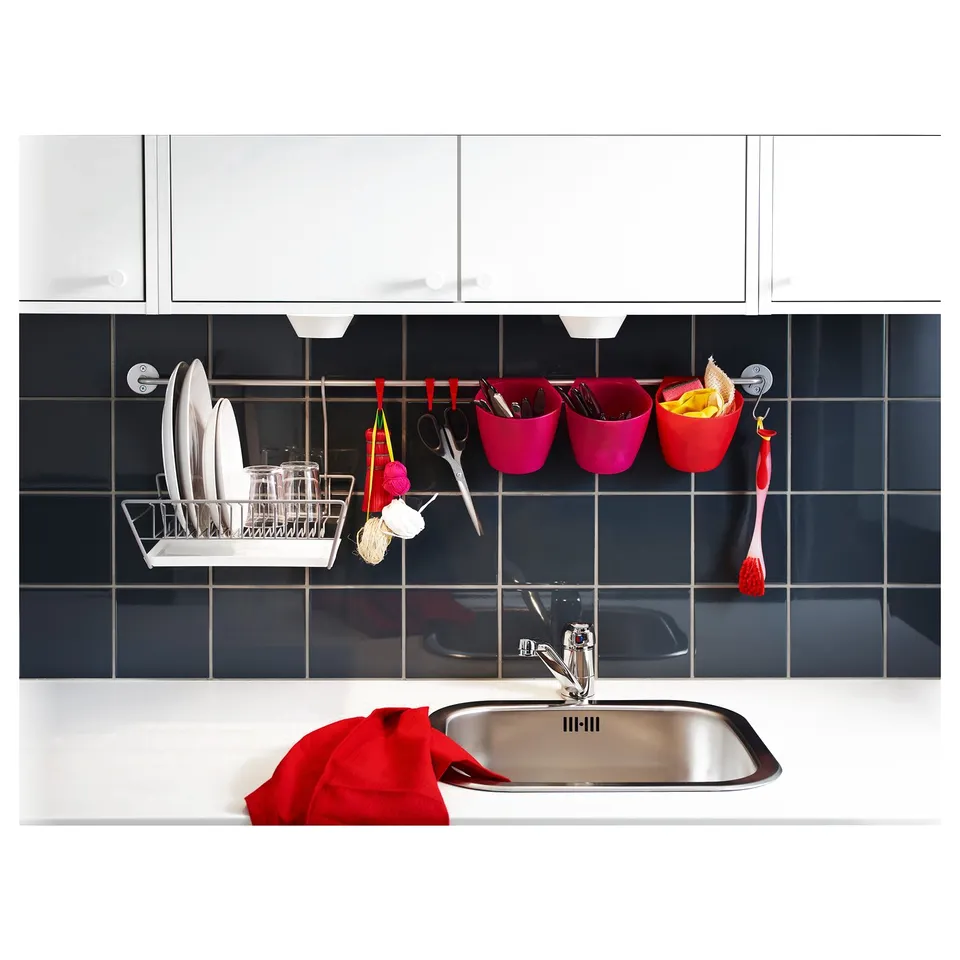 Modern kitchen sink in stainless steel. White countertop, red cloth, pink and red containers, utensils, and silverware holder against dark blue tiles.