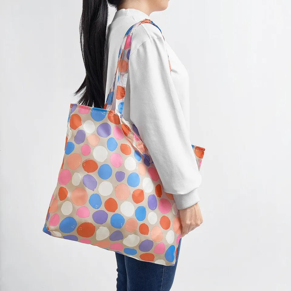 A person carrying a bright polka dot shoulder bag. The bag is foldable, lightweight, and can be used for shopping or travelling.