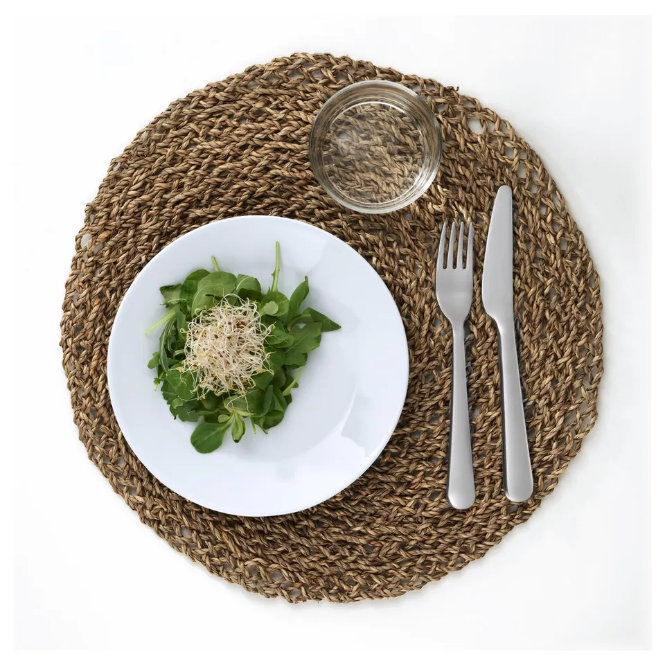 A table setting with a woven place mat featuring a white plate of green salad, a glass, and cutlery.