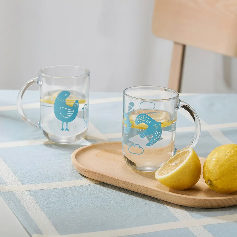 Two cups with blue bird designs and handles hold drinks on a wooden tray, accompanied by lemons on a blue checked tablecloth.