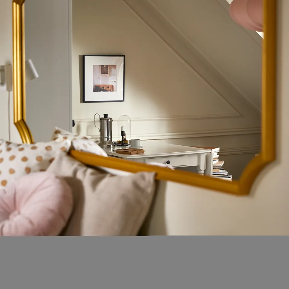 A gold-framed mirror hangs on a wall, reflecting a bedside table with a lamp, picture, and neatly made bed with pink polka dot pillows.