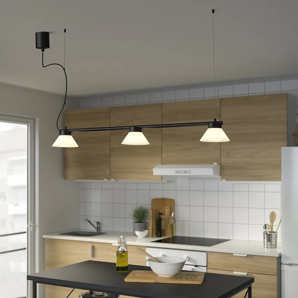 Modern kitchen with SAMBORD black pendant lamp over island. Lamp has three cones. White backsplash, wooden cabinets, and olive oil on counter.