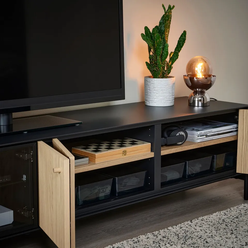 Black tv stand with bi-fold doors showing chess board, tv, plant, and lamp; glass doors, cable management.