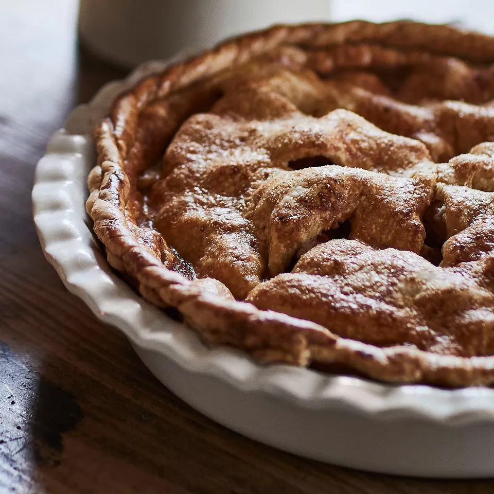 Close-up of a freshly baked apple pie in a sturdy, textured pie dish, dusted with icing sugar, sitting on a rustic table.