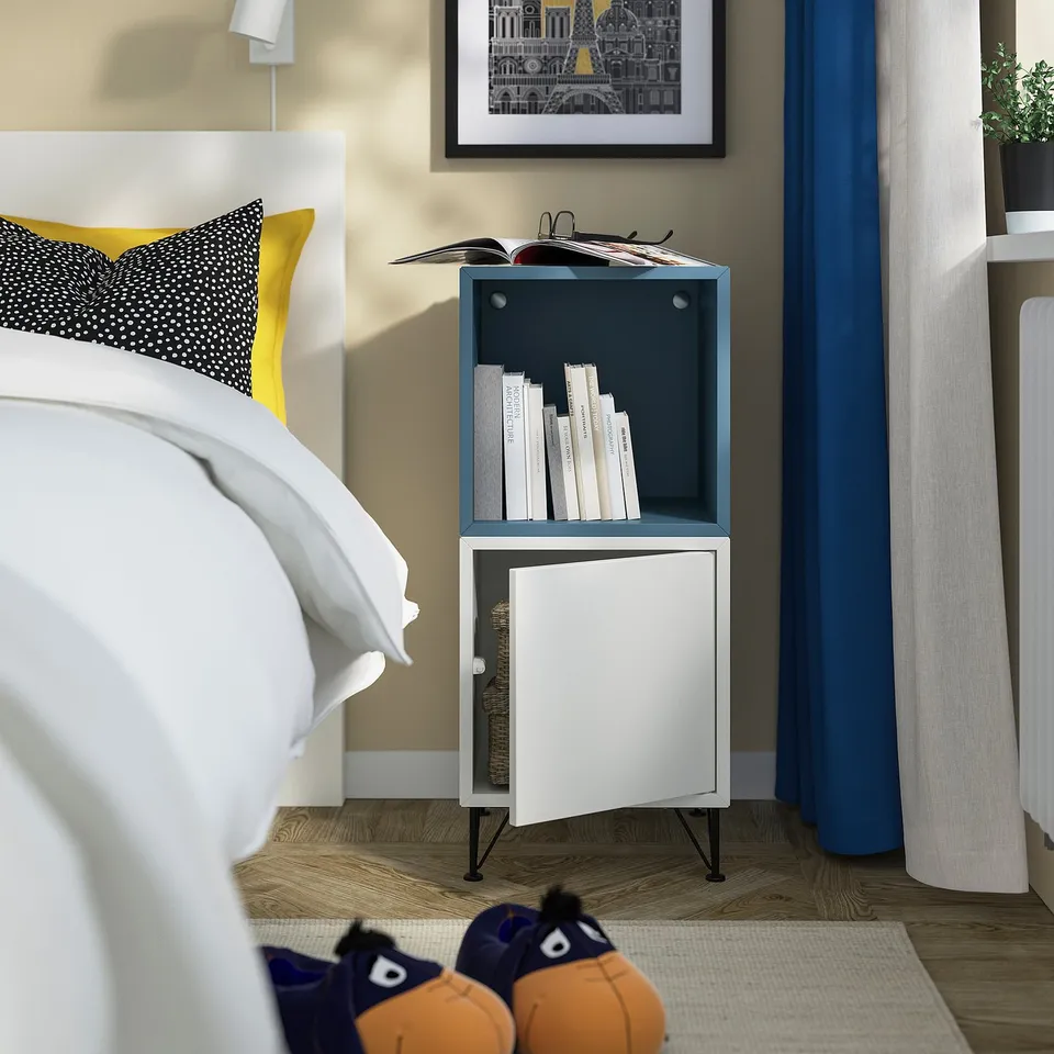 Bedroom with white bed, bright pillows, narrow bookshelf: EKET, holding books, near window with blue curtain, white rug, blue slippers.