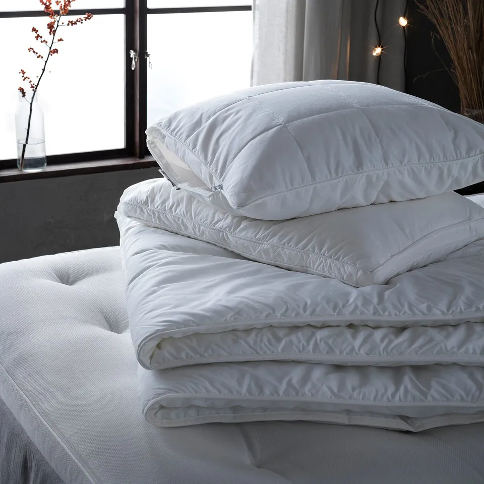 Folded white pillows with double piping on bed, natural light from window, minimalist decor.