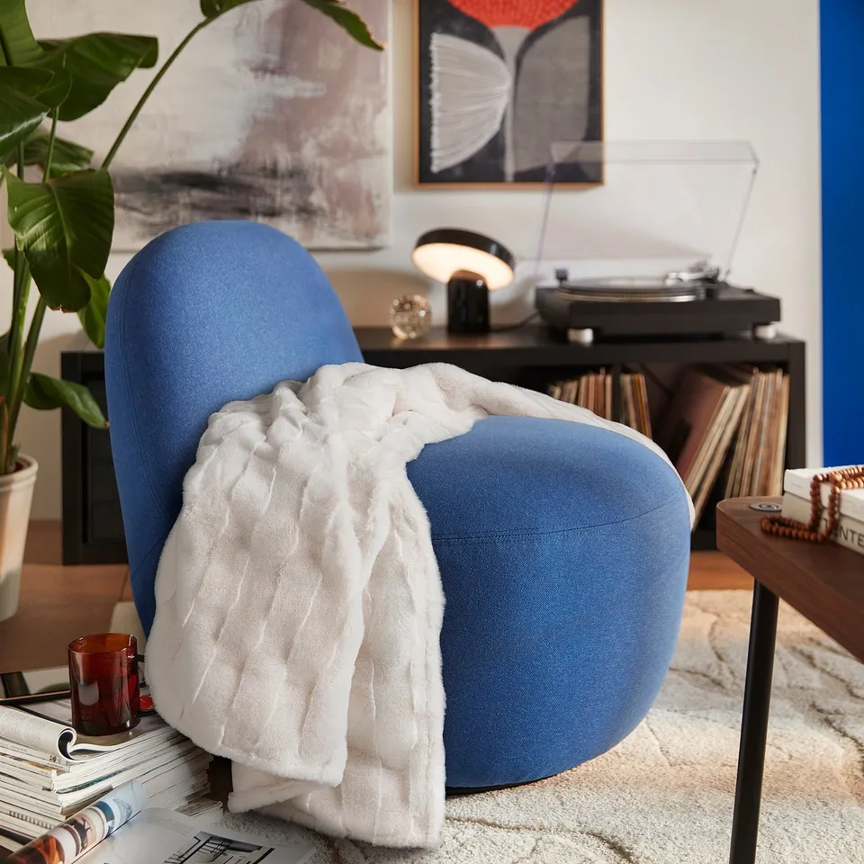 Blue, rounded TORPSPIREA chair with a white, wavy-textured throw blanket. Near a side table with records, a lamp, and a plant.