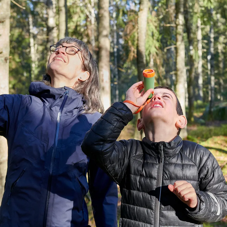 Adult and child observe trees. Child uses binoculars, adult smiles.