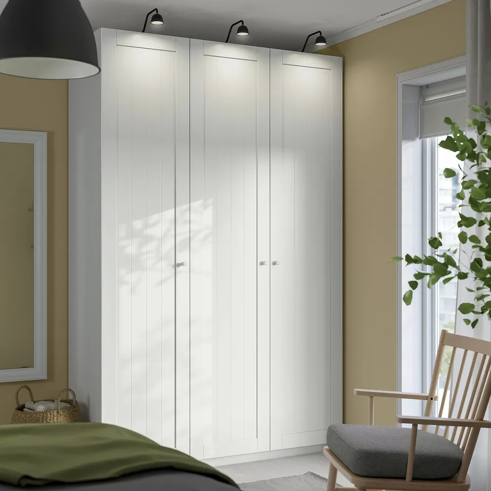 White PAX wardrobe, tall & sleek, beside window, next to wooden rocking chair. Tall, minimalist design in calm bedroom.