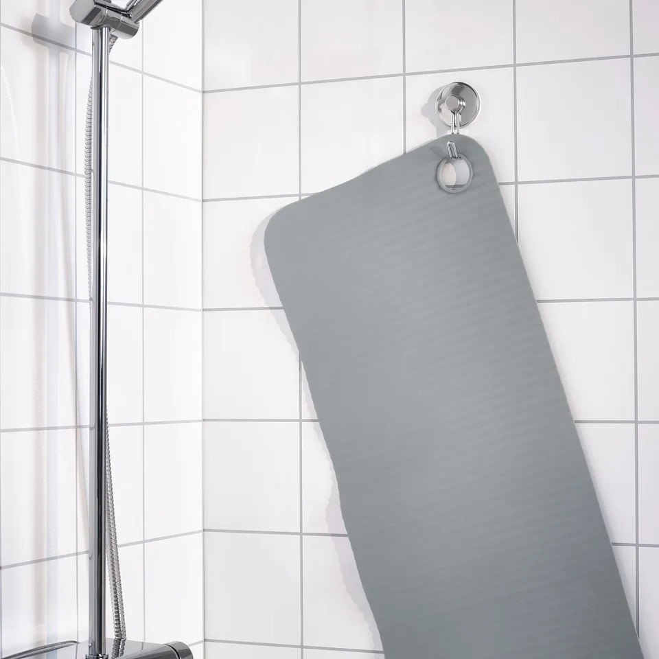 A grey bath mat with suction hooks securely hangs on tiled wall, designed to stay in place and prevent slipping.