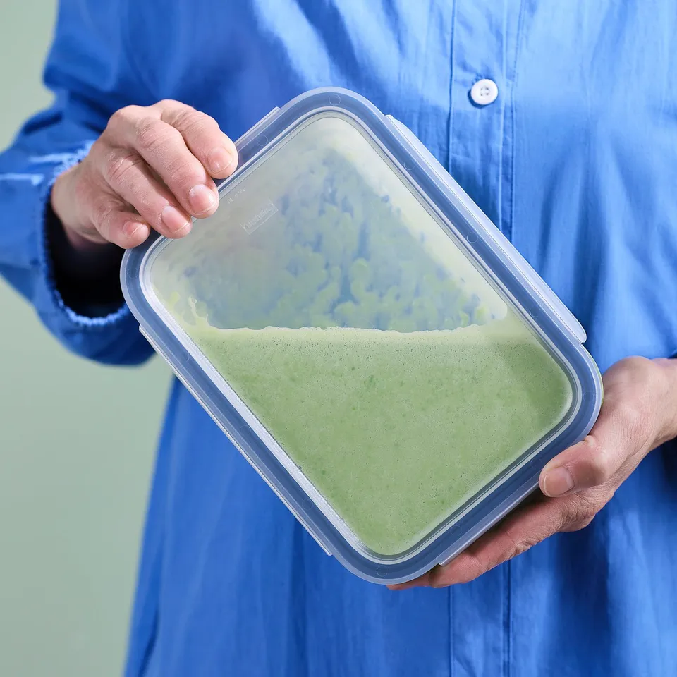 Person holding a clear plastic food container with green liquid, likely soup or sauce. The container has a snap-lock lid and is made by IKEA.