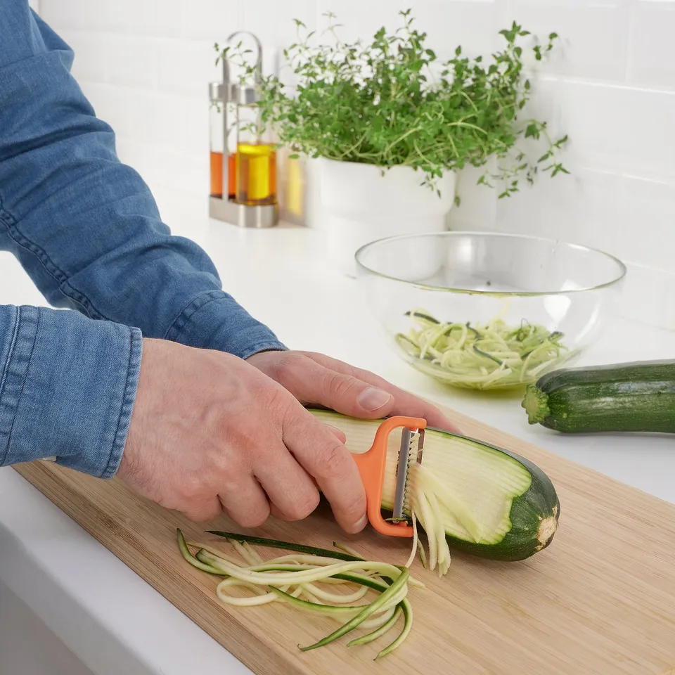Person peels courgette into strips using orange UPPFYLLD vegetable cutter on wooden board. Bowl of peeled courgette and herbs in background.
