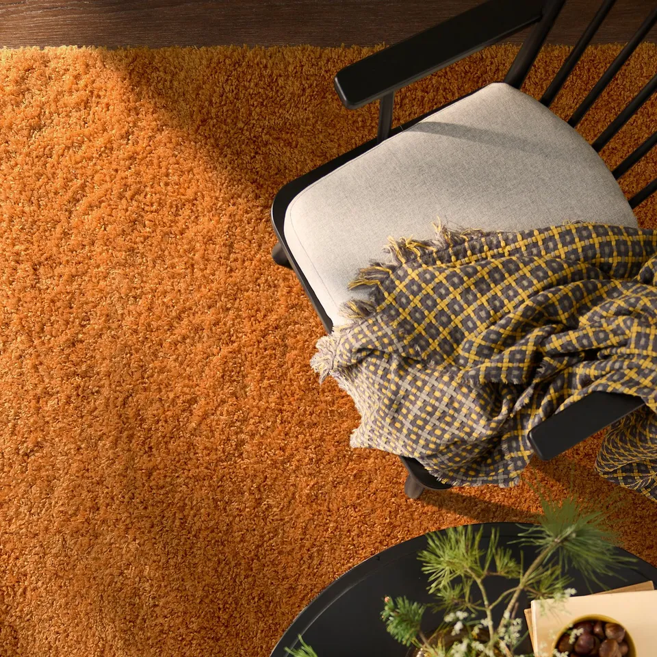 Brown rug with high pile, partially hidden by black chair with grey cushion and yellow-blue cloth. Soft, contrasts with wood floor.