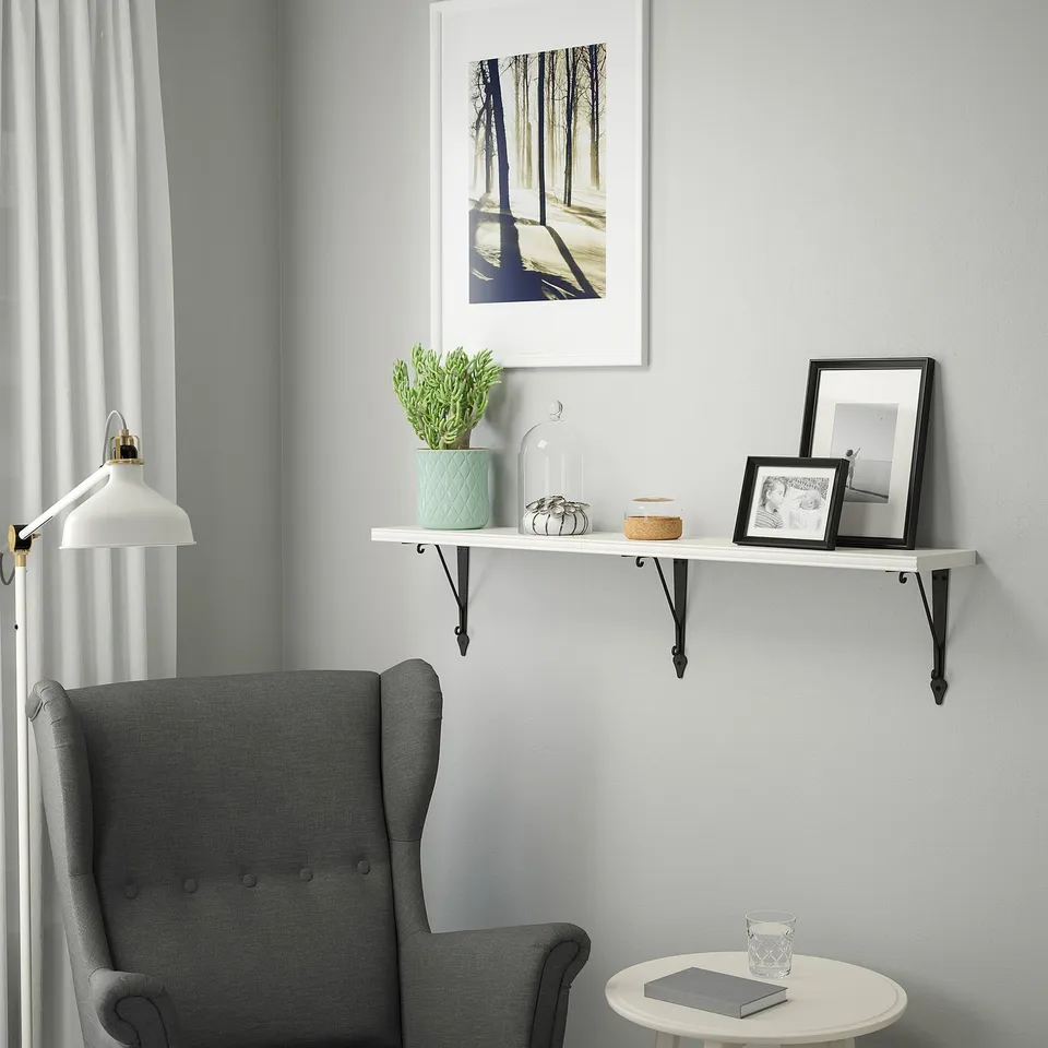 A grey armchair beside a wall shelf holding a framed photo, plant, candle holder, and other small items. Above the shelf is a forest picture.