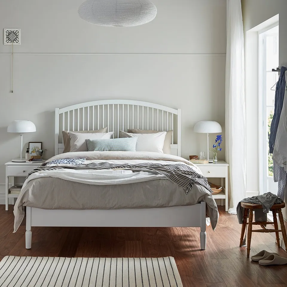 Bright, white wooden bed with striped linens and pillows, flanked by lamps, against a light wall with a textured ceiling light.