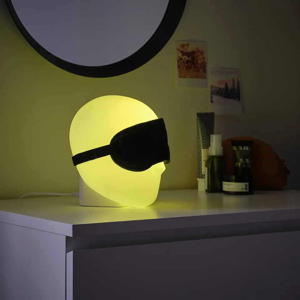 ISKÄRNA lamp with yellow light, cylindrical shape, and black eye mask design on a table with decor items.