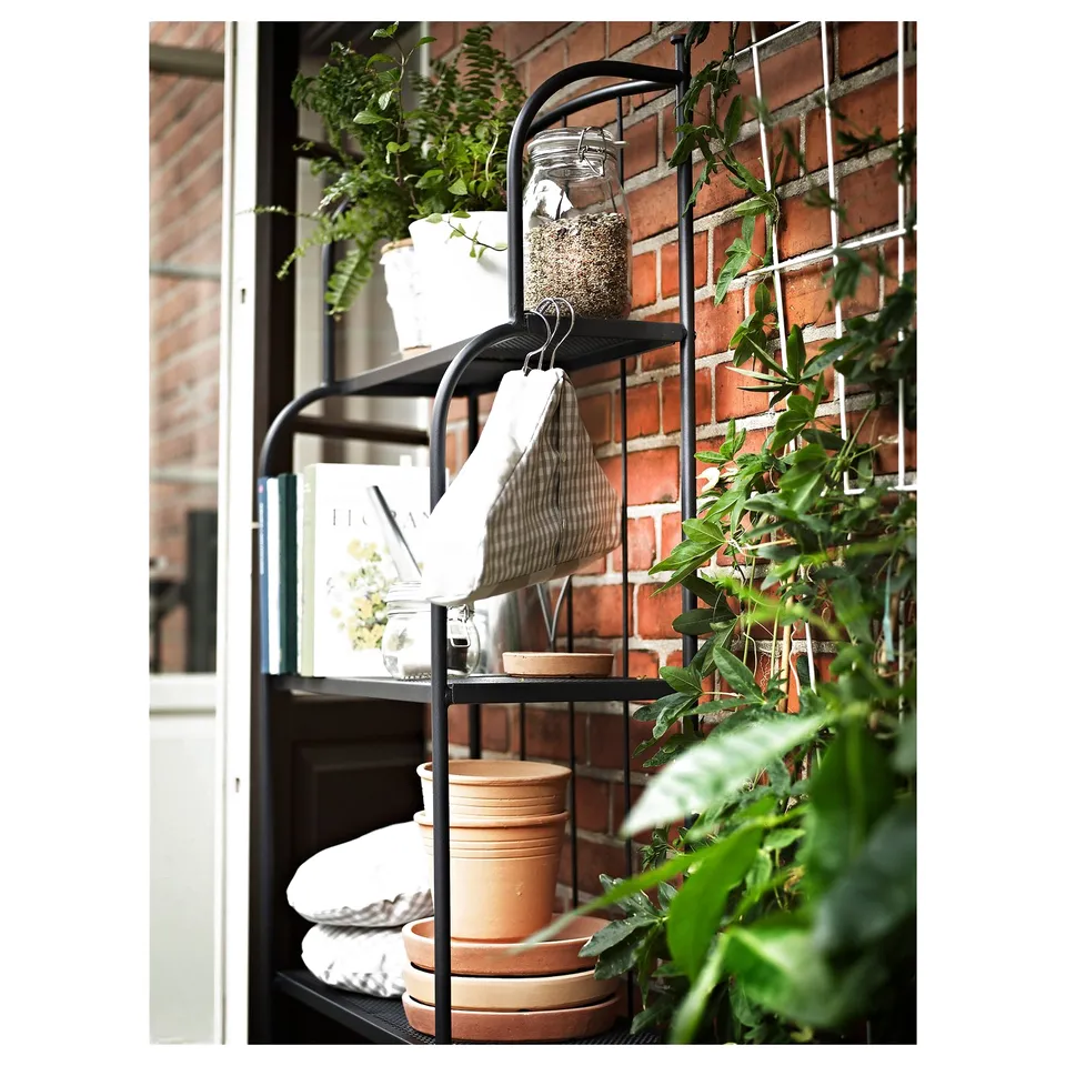 Indoor black metal plant stand with potted greenery, jars, and linens against brick wall.
