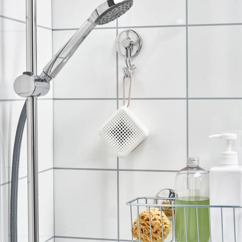 Shower setup with a white square speaker hanging from a metallic hook, secured by suction cups. Nearby, a wire basket holds items.