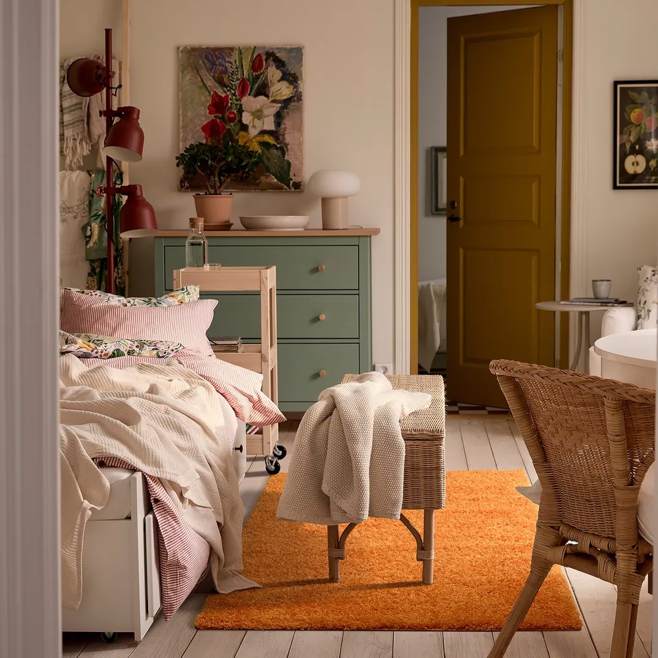 Bright bedroom with an orange arende rug, wicker chair, white bedding, green dresser, and floral art. Soft, thick rug contrasts cosy decor.