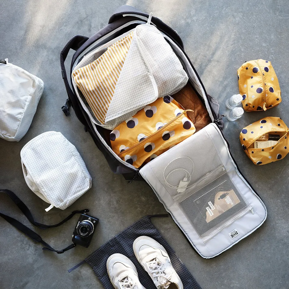 Open backpack with folded clothes and hairs accessories. Yellow polka and striped pouches, white trainers, camera, and grey chequered pants nearby.