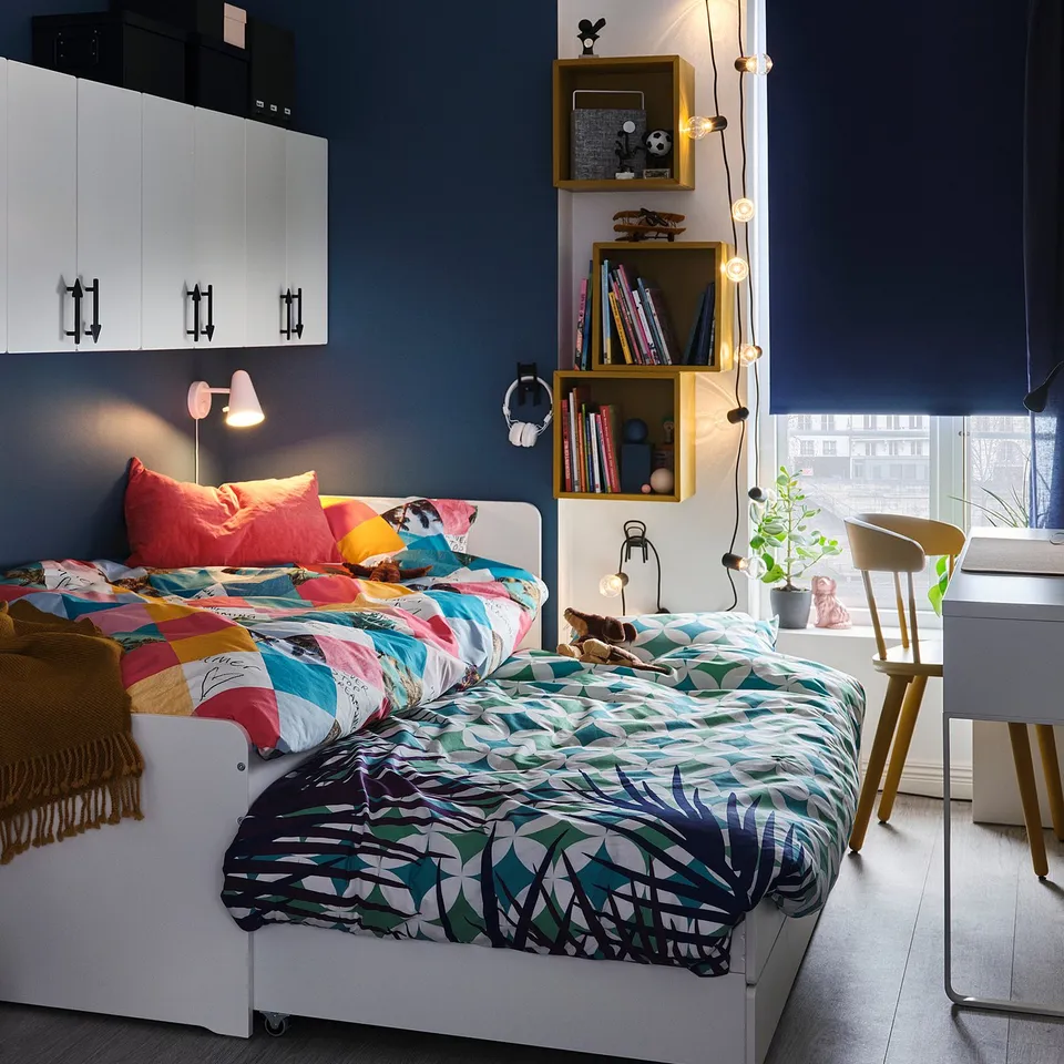 Vibrant kids room with blue walls, two white beds with colourful linens, wooden shelves, and a desk by the window.