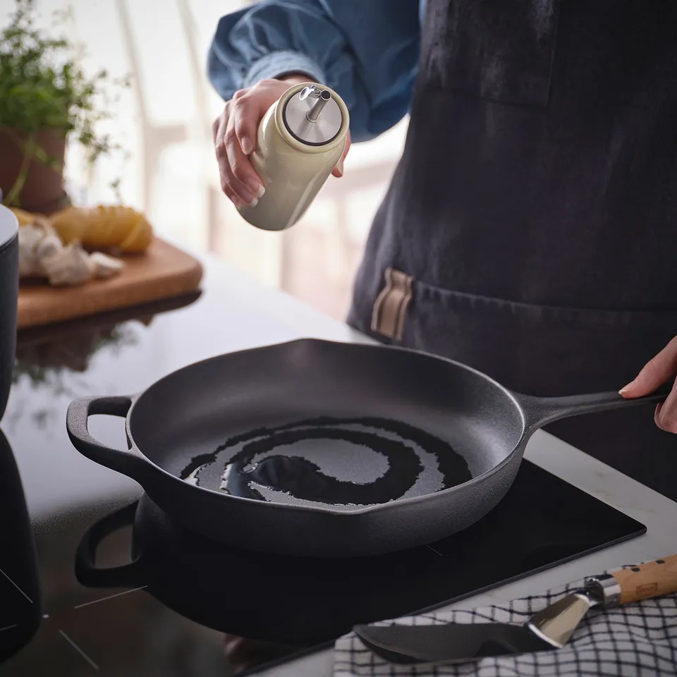 Person seasoning pan with oil from BRUGDHAJ bottle. Pan sits on stove with ingredients nearby.