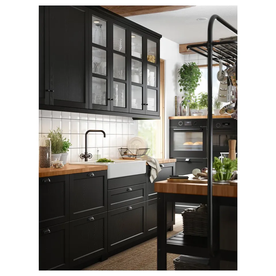 Modern kitchen with black cabinets, white countertop, farm sink, wooden counter, potted plants, hanging rack with tools, and a large window.