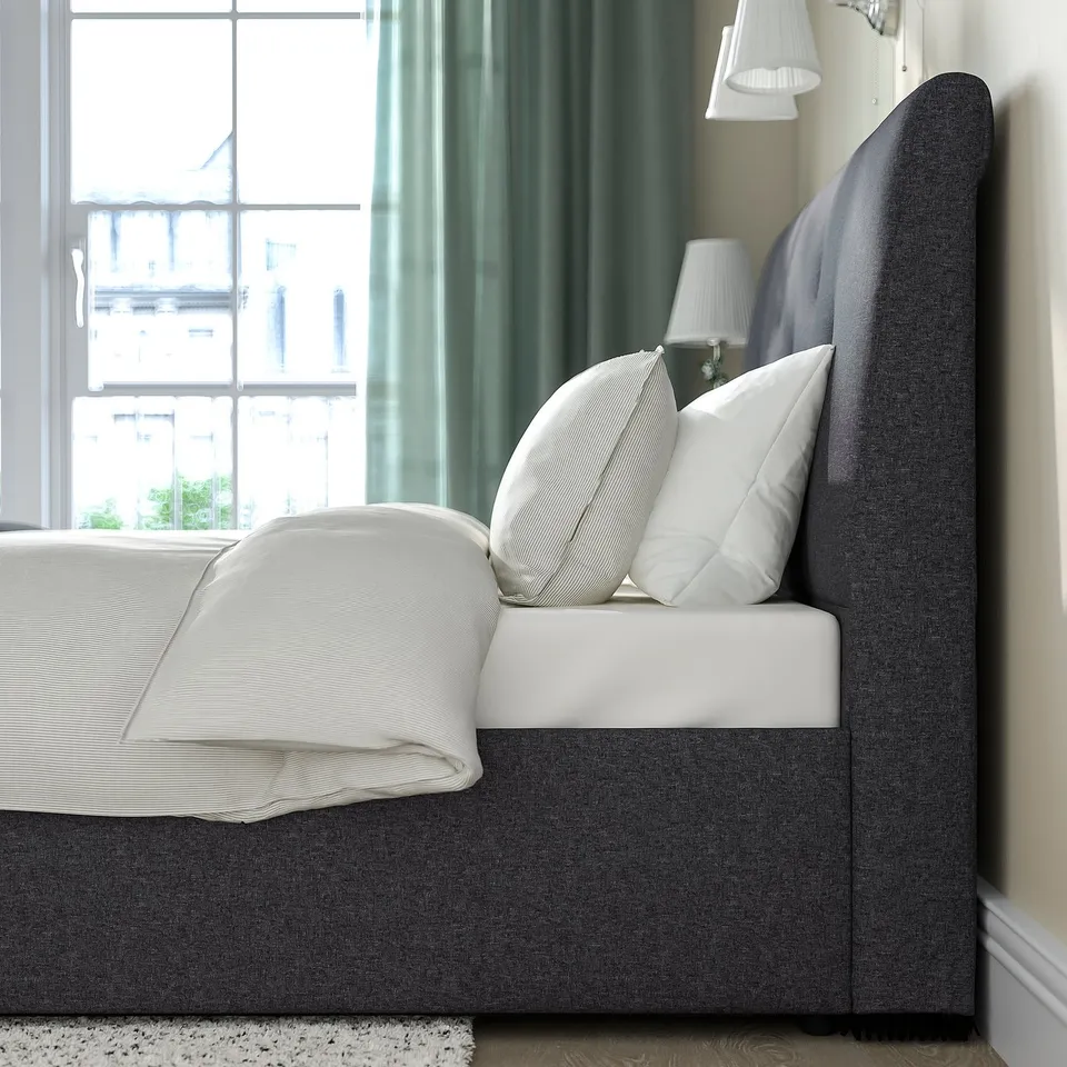 Upholstered bed with sloped headboard, white pillows, and blanket. Grey fabric with integrated lift for under-bed storage.