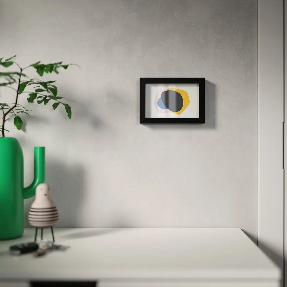 Abstract art in black frame on grey wall, above white table with green vase and plant.