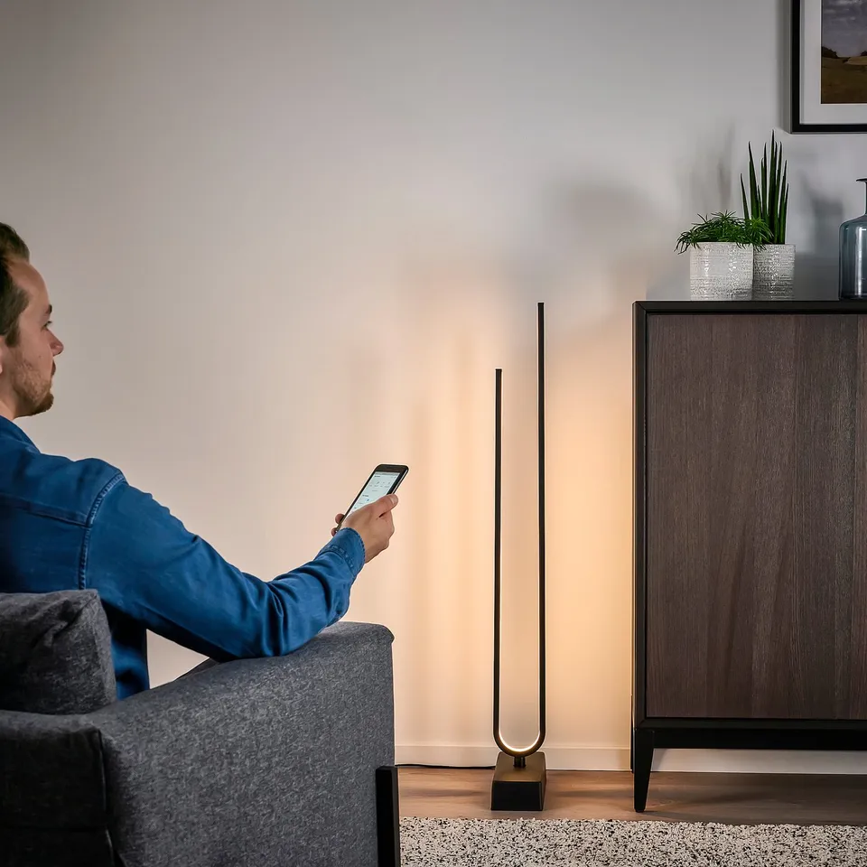 Person in blue shirt sits on grey couch, using smartphone beside modern floor lamp with dynamic lighting.