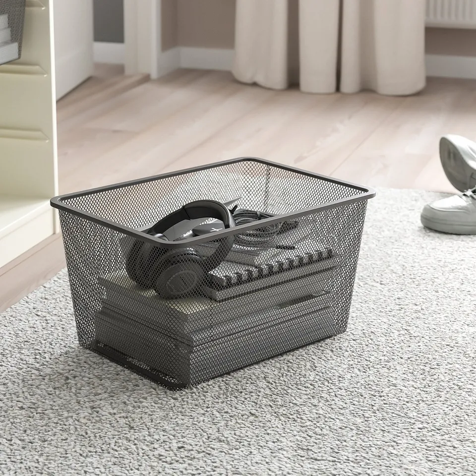 Black mesh storage basket for organising items, placed on a grey carpet in a bedroom.