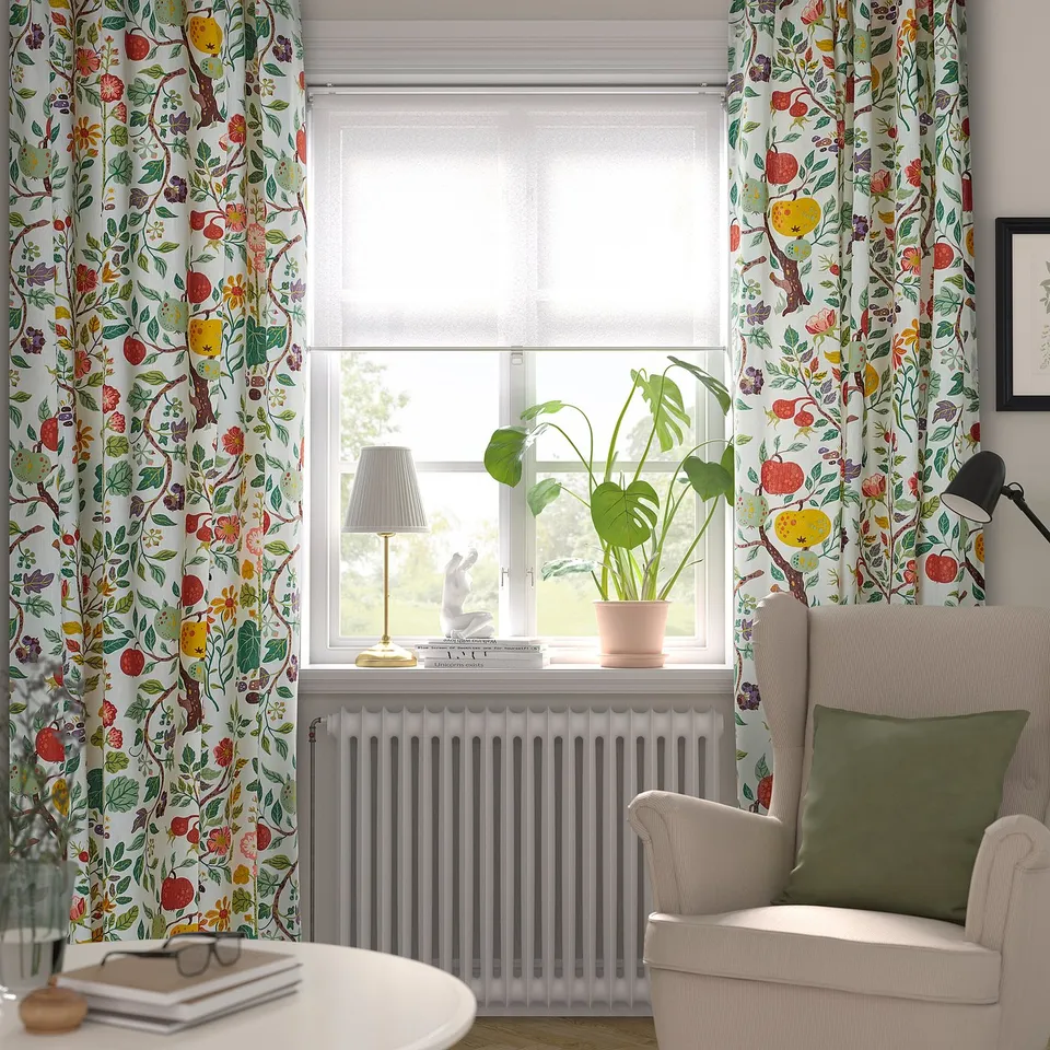 Window with floral curtains, cosy chair, plant, and books.