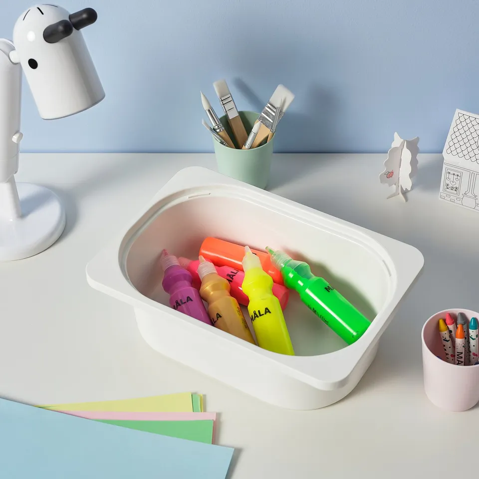 White storage box filled with colourful highlighters sits on a light desk, surrounded by art supplies.