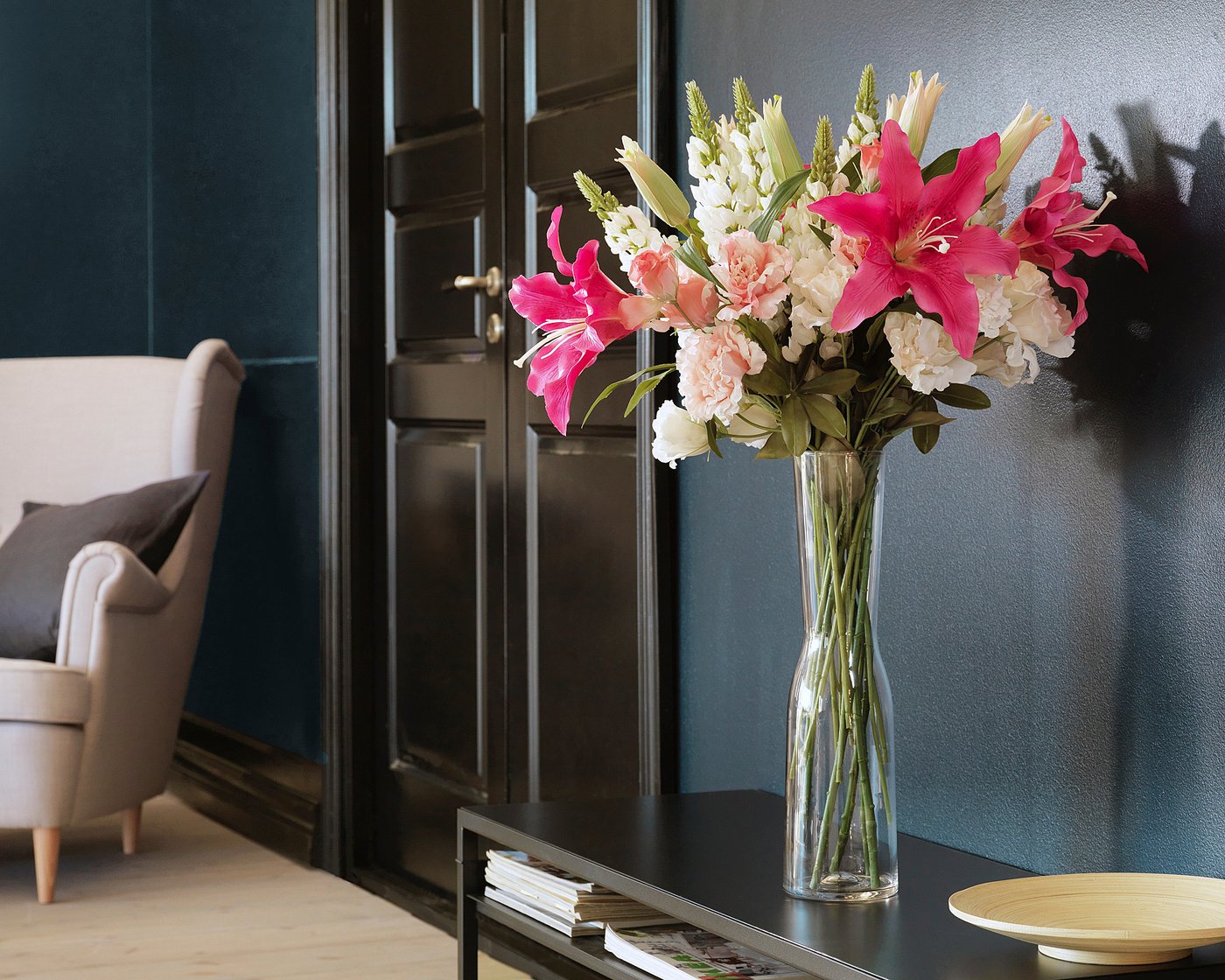Vase with pink lilies, white flowers on dark table, plush chair, dark teal wall.
