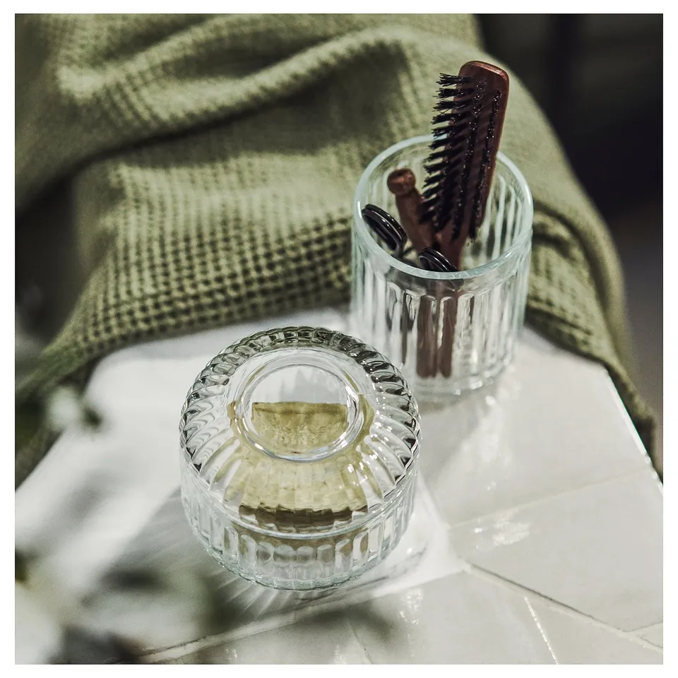 Clear glass bathroom accessories, one with lid, contain items. Sleek, ribbed design, sits on green cloth.