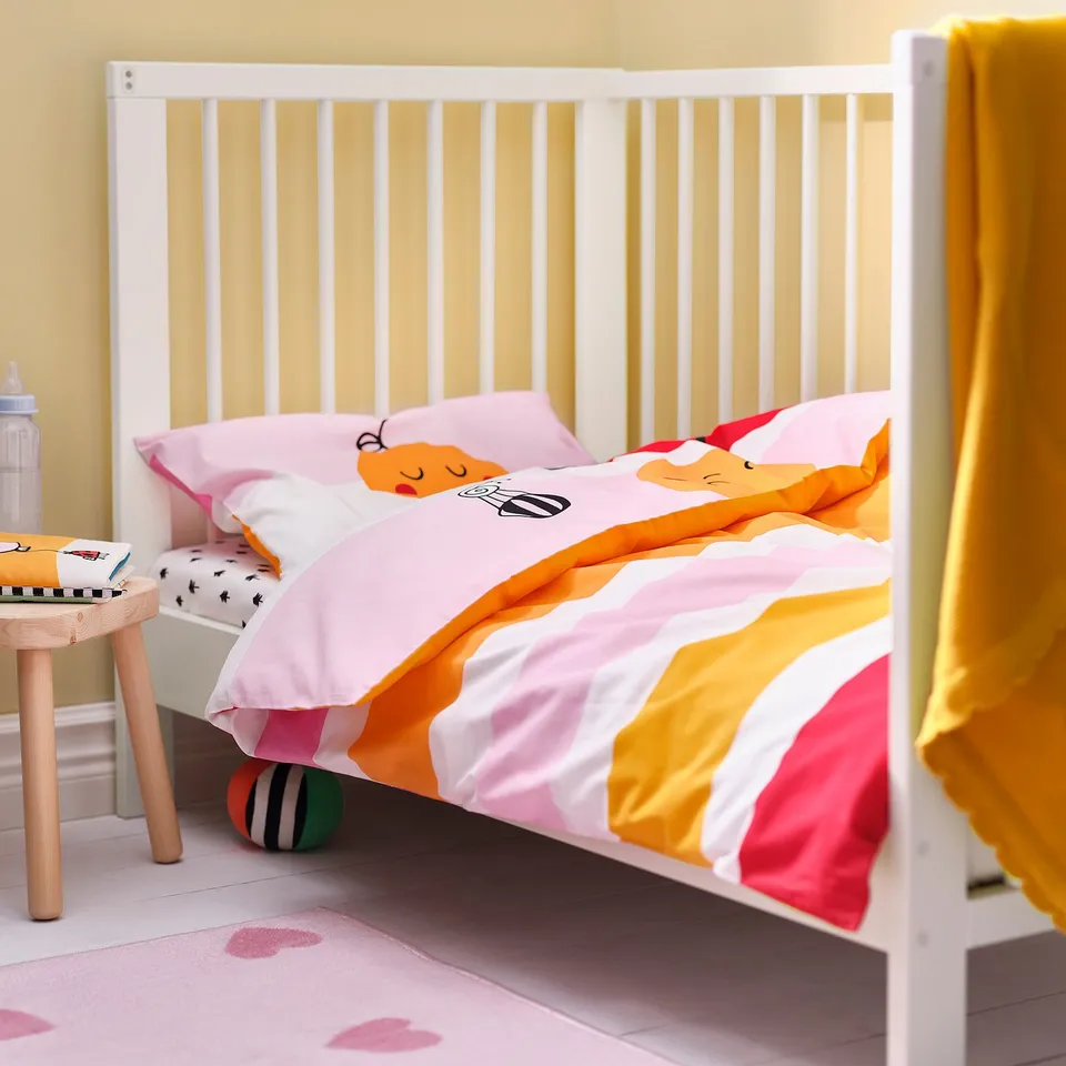 Colourful baby crib with cartoon duvet, yellow curtain, stool, bottle & book in bright room.