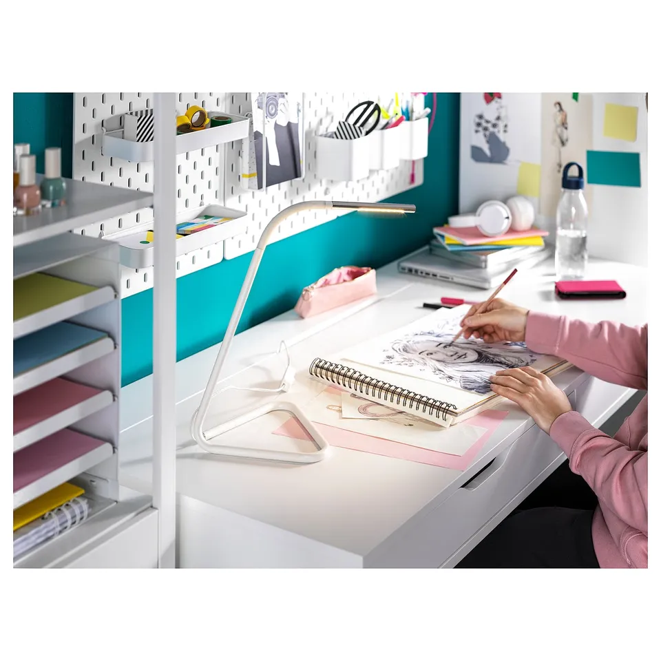 White ALEX desk with built-in drawers. Person drawing on paper; stacks of books, pink accessories, and a water bottle on the desk.