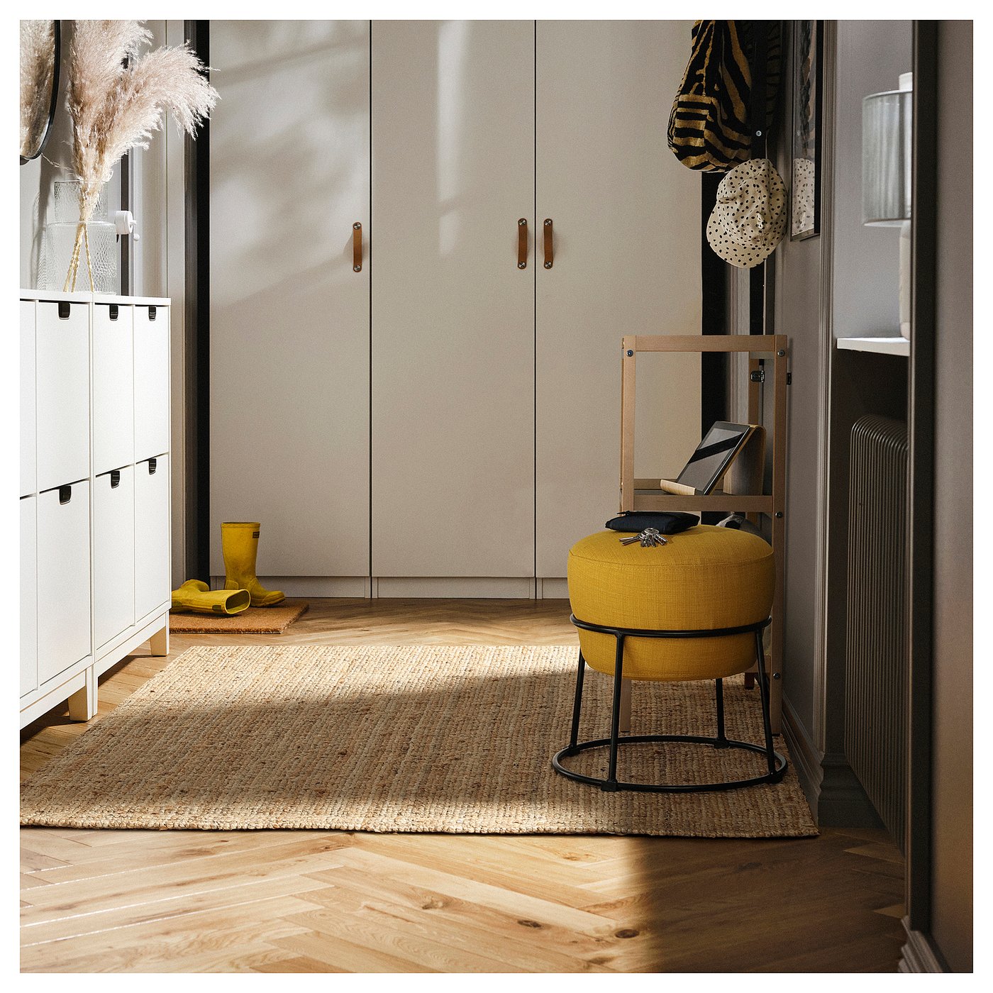 Modern room with wooden floors, off-white closets, yellow pouffe on jute rug, boots, radiator, books, dining table with feathers.