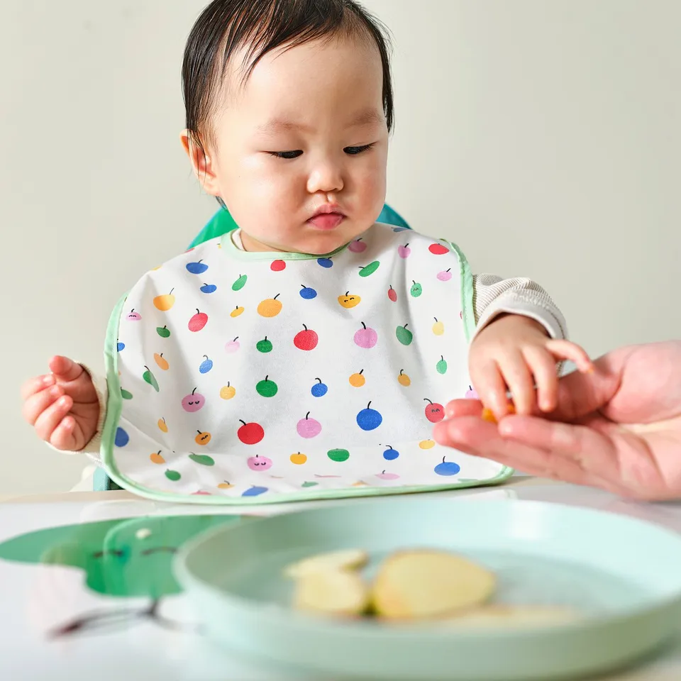 Baby in fruit bib sits at table, adult offers food.