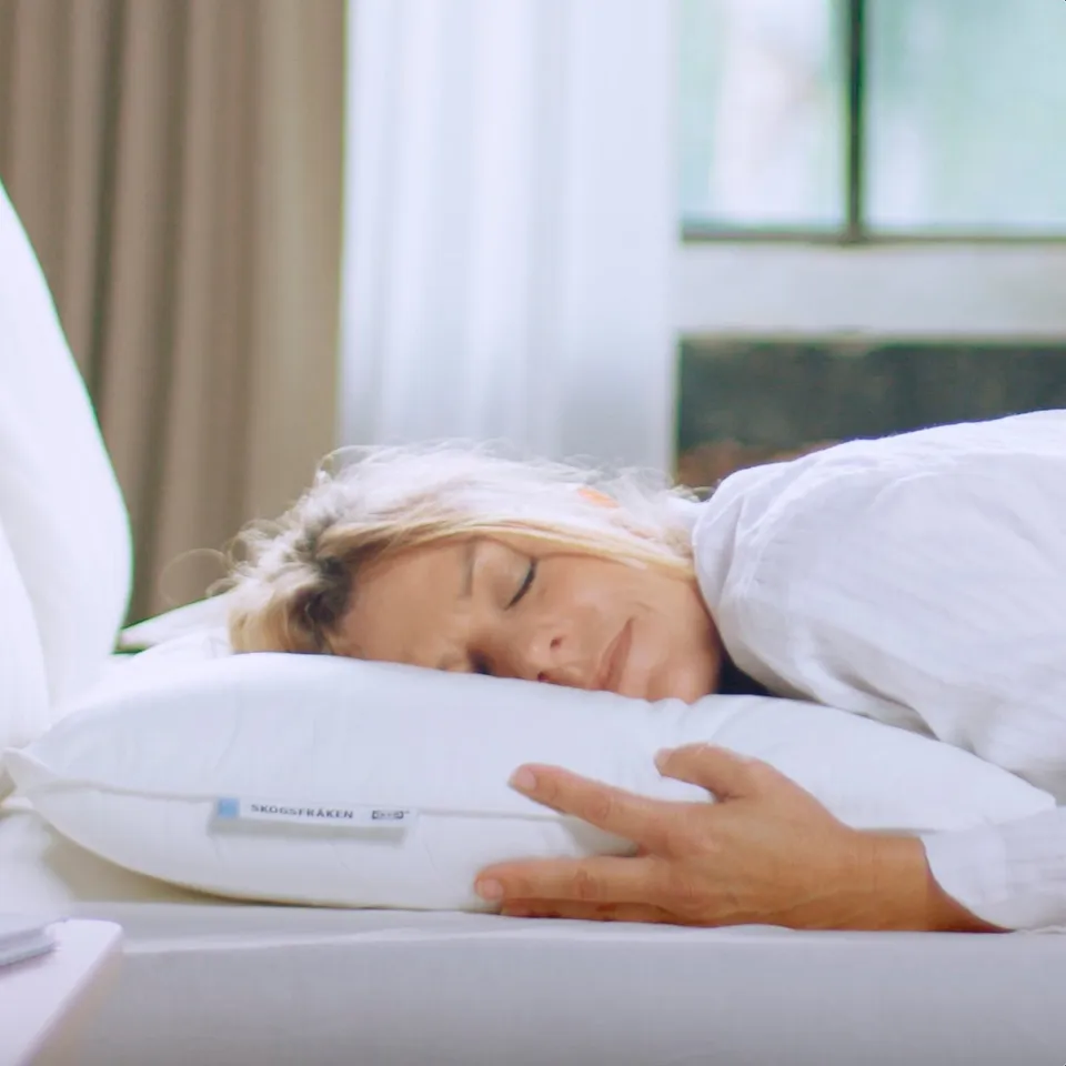 A person sleeps peacefully on a white pillow labelled SKOGSFRÄKEN on a bed with white sheets.