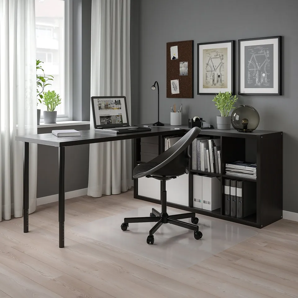 Home office setup with KALLAX unit. Modern black desk, chair, and shelf. Laptop, books, and plants arranged. Large window on left, grey walls.