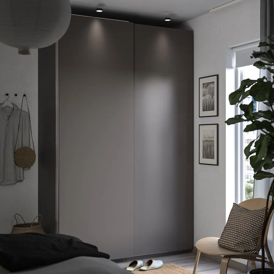 Modern bedroom with PAX wardrobe, sliding dark doors, hanging clothes, wooden chair, plant, and framed pictures.