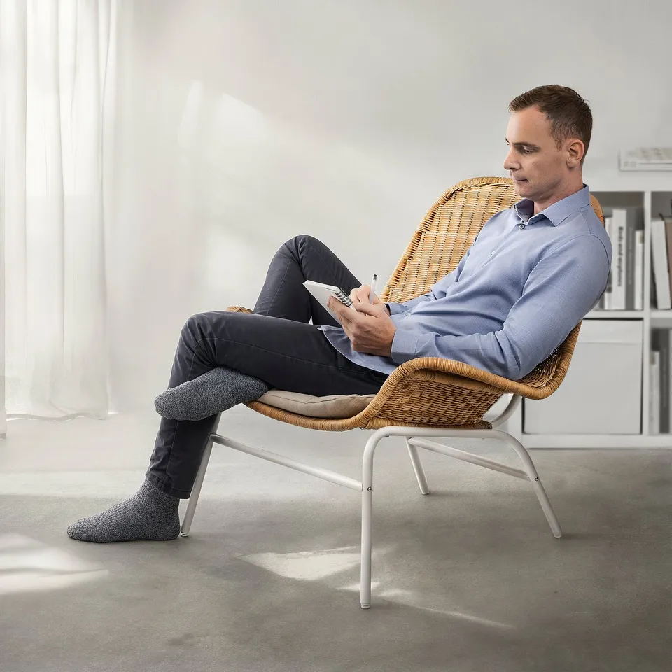 Person sitting in wicker FRYKSÅS chair, holding paper, in bright room. Chair is compact, smaller than rooms bookshelf.