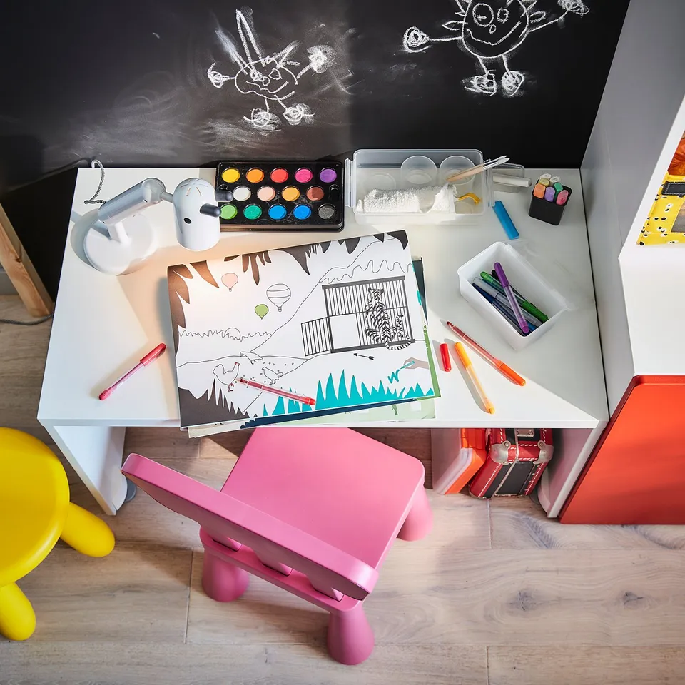 Childs craft table with pink chair, adjustable white lamp, coloured pens, paints, and drawing paper on blackboard wall.