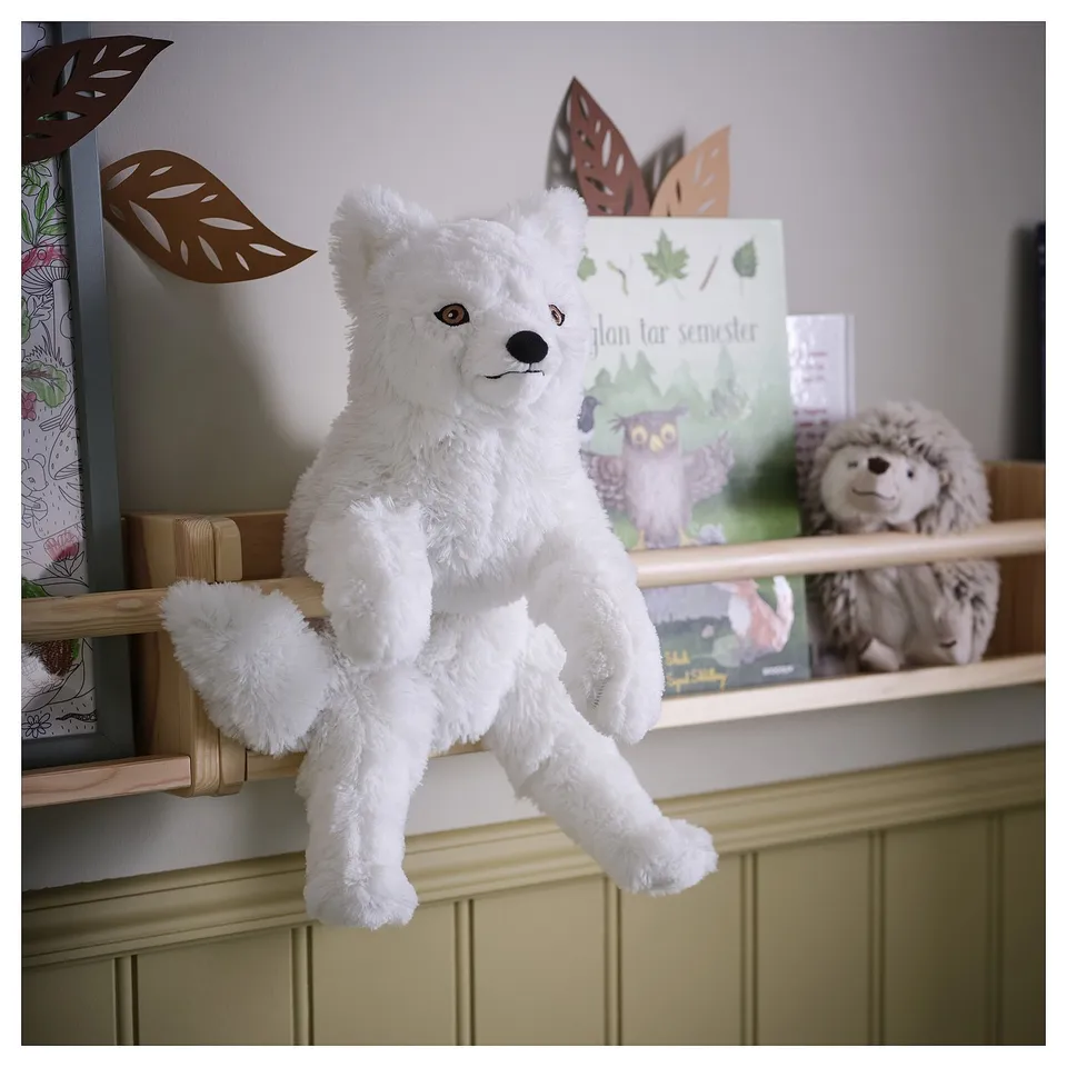 White plush arctic fox toy sits on wooden shelf with beige hedgehog, book titled plan tar semester, and decorative leaves.
