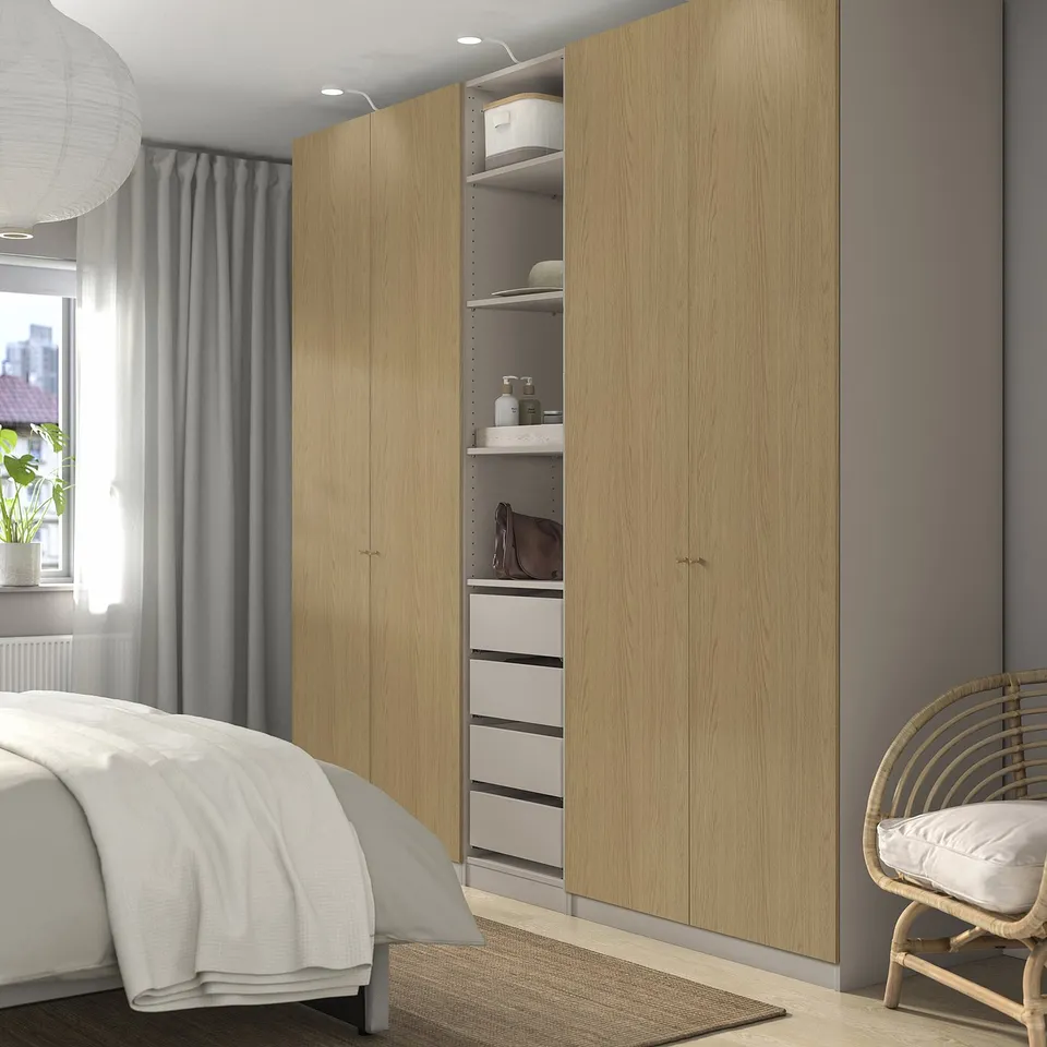 Spacious bedroom with light wood PAX wardrobe, open section, white rug, wicker chair, window, grey curtains, and bed with white linens.
