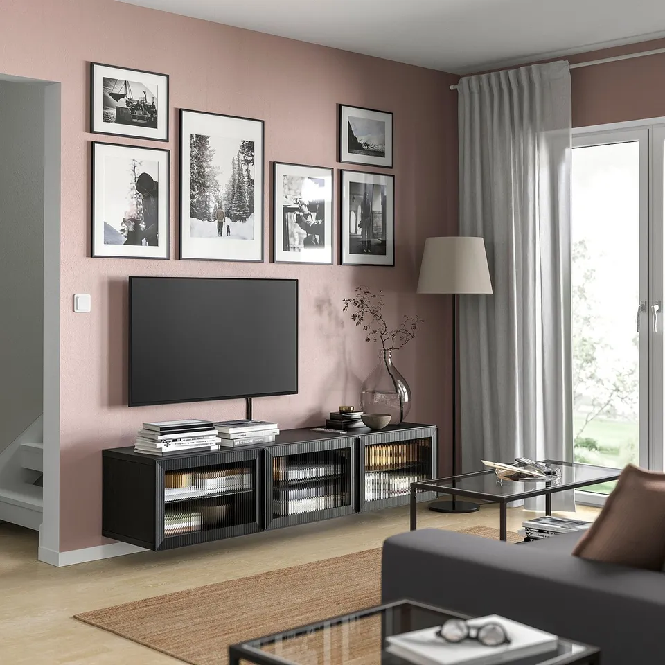 Modern living room with BESTÅ tv bench, flat screen, and decor. Pink walls feature black photo frames, grey curtain, wooden floor.
