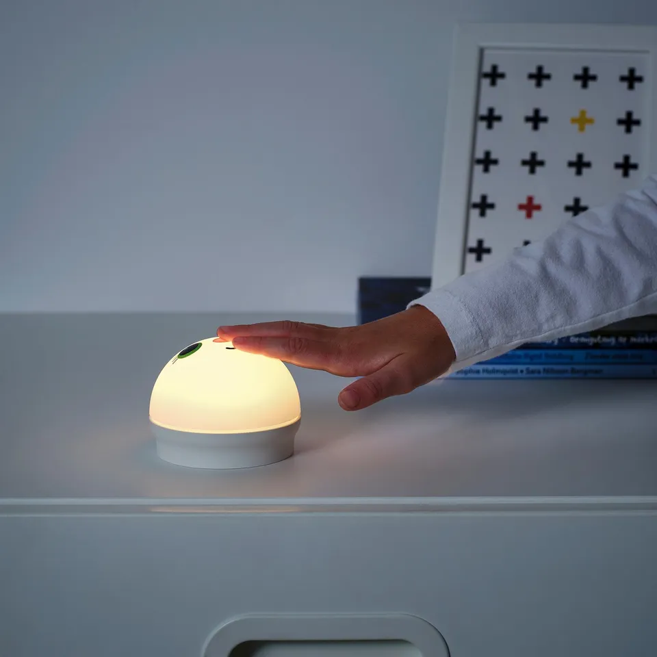 A person turns on a small, round nightlight, KORNSNÖ, which emits a soft glow.